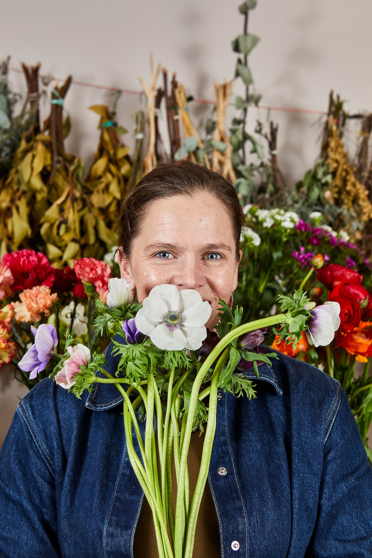 A smiling woman with short brown hair holds a large bouquet of white anemone flowers in front of her face. She is wearing a blue denim jacket, and a colorful, rustic dried flower arrangement hangs on a string in the background against a neutral wall.